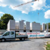Neubau-Baustelle mit weißen Betonwänden und Emsbau-Firmenfahrzeug vor Bauzaun mit Werbeplakat