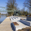 Moderne Hafenanlage mit Betontreppe zum Wasser, Holzbänken und Stahlbrücke im Hintergrund bei blauem Himmel