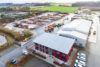 Luftaufnahme des modernen Firmenstandorts mit rotem Verwaltungsgebäude, Lagerhallen und Baustoffhof in ländlicher Umgebung